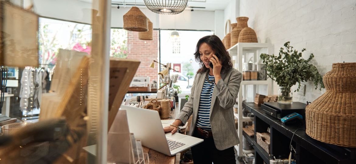 Smiling young Asian woman standing behind a counter in her stylish boutique working on a laptop and talking on a cellphone