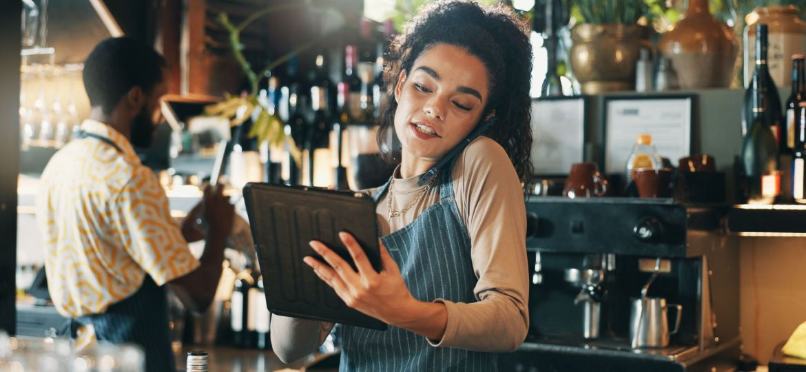 A woman taking a phone call and using a tablet behind the counter at a cafe.